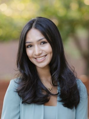 A woman with long dark brown hair smiles, wearing a teal blazer and shirt in front of an outdoor background with trees