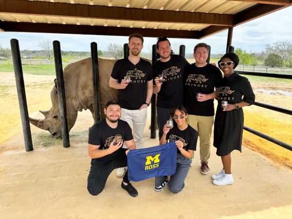 Students stand in front of a rhino in a cage holding a Michigan Ross flag