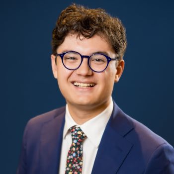 A student with short brown hair, glasses, and a blue suit with white dress shirt and flowery tie