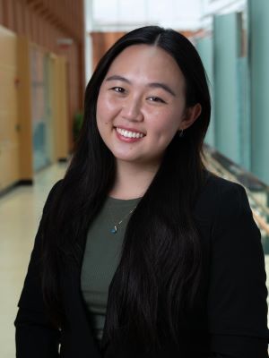 A woman with long black hair smiling, wearing a black blazer and green shirt with the Ross Winter Garden in the background