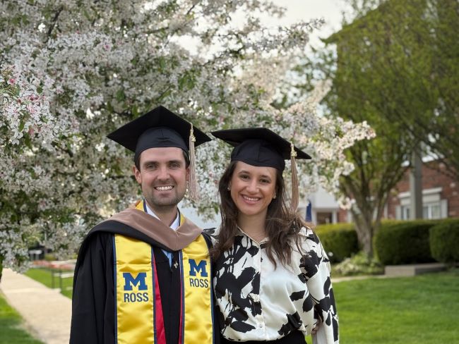 A couple smiles, wearing graduation caps, on a street with flowering trees in the background.