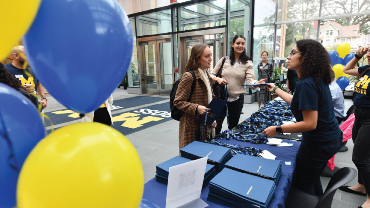 Prospective students interacting with an admissons representative at a Michigan Ross table in a building. There are blue and yellow balloons in the left of the image.