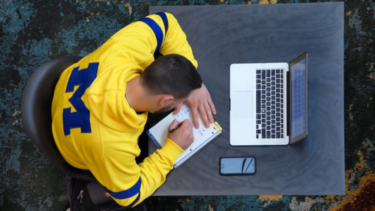 Man wearing a maize shirt with a navy blue block M on the back sitting in front of a computer and writing in a notebook.