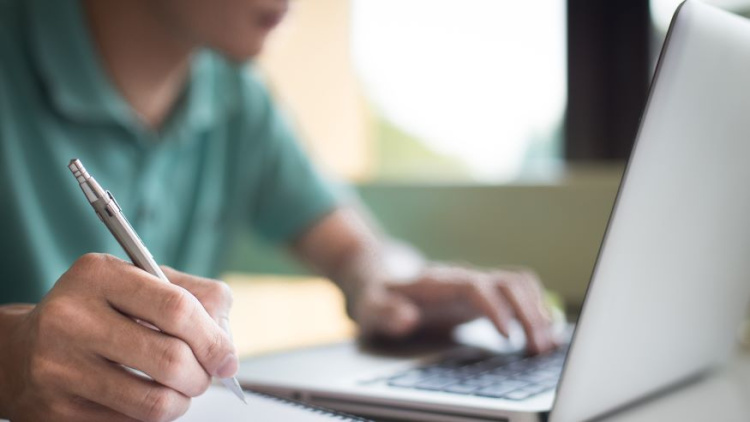 Photo of a hand on a laptop computer and one hand holding a pen and writing in a notebook. You can't see the man's face but he is wearing a teal green shirt. 