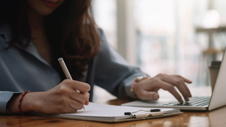 A woman types on her computer at a table while writing notes on a paper on a clipboard with a pen
