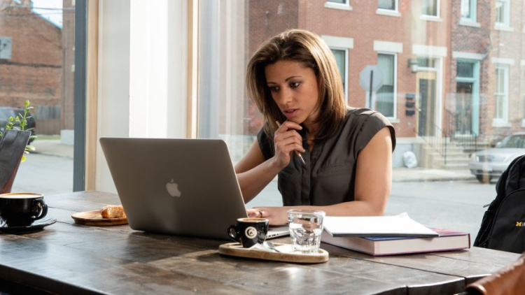 Photo of a woman sitting at table looking at her laptop holding a pencil in her hand. She has medium length dark hair and is wearing a brown short sleeve shirt. 