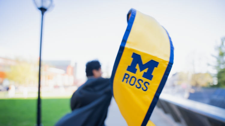 Photo of the graduation shawl for the Ross School of Business. It is maize and blue with a block M and Ross written underneath it. There is a student in the background who is holding it up. 