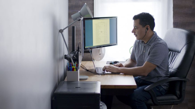 Photo of a man sitting at a computer in his home looking at the screen. He is wearing a light blue shirt and has dark hair. 