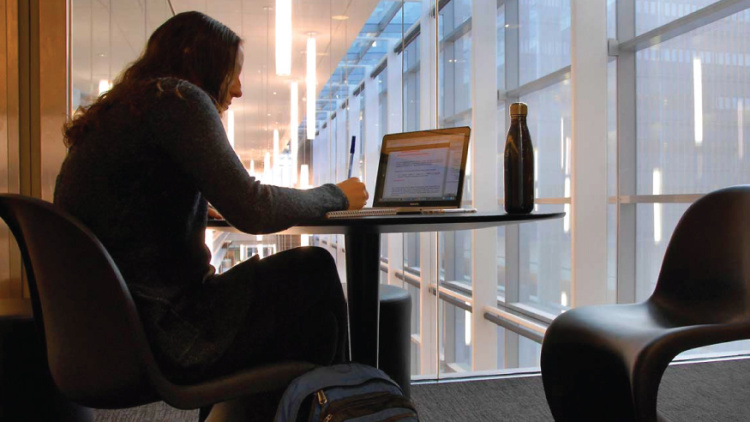 A person with long hair writes in a notebook while looking at a laptop screen on a table.