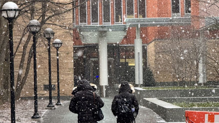 Photo of two Michigan Ross students walking into the building. You only see their backs and they are both wearing black coats. There is snow falling all around. 