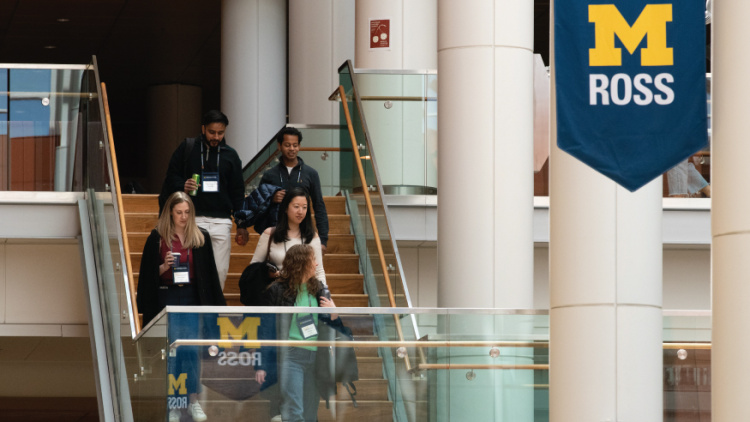 A group of students descends the staircase in the Michigan Ross Winter Garden under a blue ballner with the Block M and "Ross" on it.