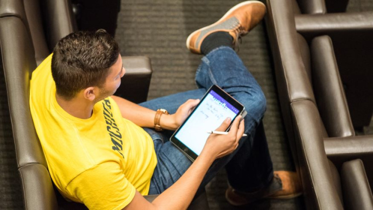 Photo of a man wearing a yellow Michigan shirt sitting in a lecture hall with an ipad on his lap. He is wearing jeans and brown shoes and he has short dark hair. 