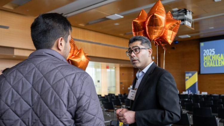 Two Michigan Ross men standing in a room for the Michigan Business Challenge. One man has his back to the camera but he has dark hair. The man who is looking at the camera has dark hair and glasses and is wearing a navy blue suit jacket. There are orange star shaped balloons in the background as well.