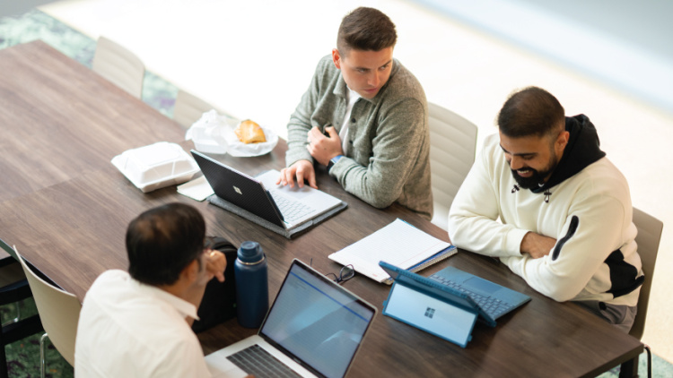 Three people sitting at a table collaborating with laptops and notebooks.
