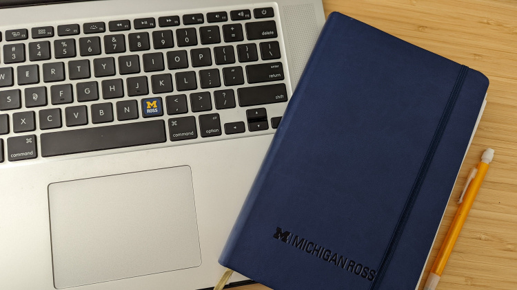 Photo of a computer keyboard next to a Michigan Ross blue notebook. There is a yellow pencil next to the notebook on the table. 