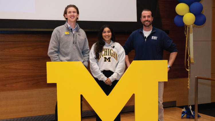 Three people stand behind a large yellow Michigan "M" logo at an indoor event.