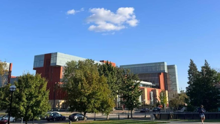Ross School of Business building is featured in the background. There are large green trees in the foreground and the sky is bright blue with one cloud that is in the shape of a block M. 