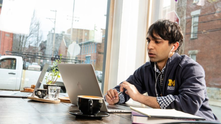 Man sitting at a table looking at his laptop. He is wearing a Michigan Ross sweatshirt and he has dark hair. There is a coffee cup next to his computer and there is a white truck outside the window he is sitting near. 