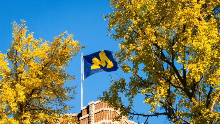 Photo of the University of Michigan block M flag flying on top of a building. It is surrounded by yellow trees and and the sky is blue with no clouds. 
