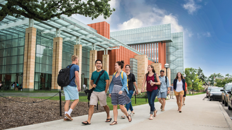 Students walking outside a modern glass-fronted campus building on a sunny day.