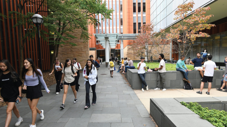 Students walking through a modern university campus courtyard with tall buildings and greenery.