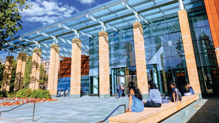 Modern glass-fronted university building with students sitting on outdoor steps on a sunny day.