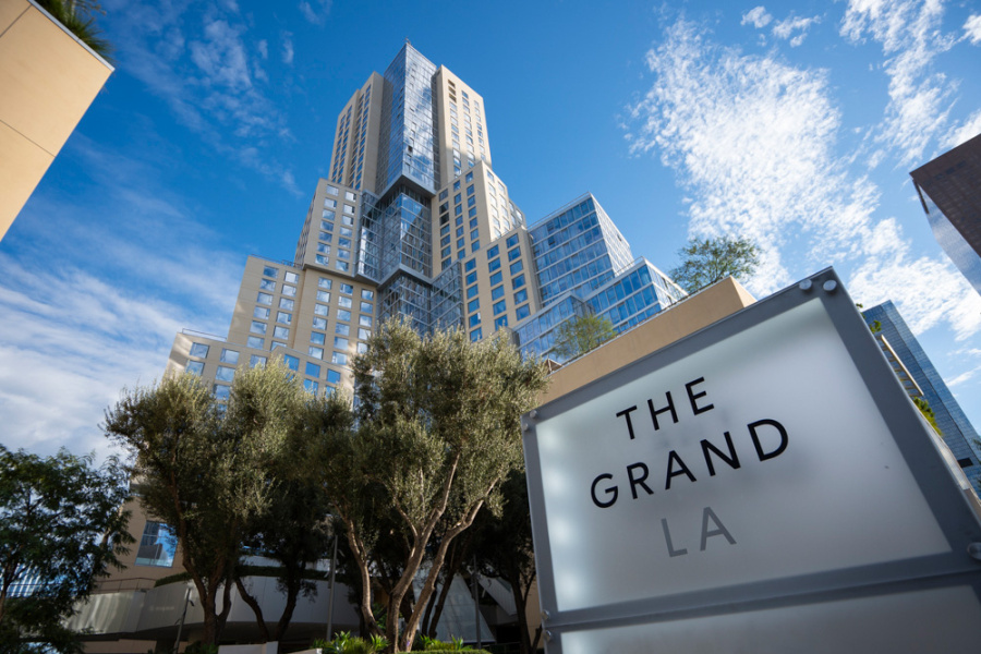 Modern, multi-tiered skyscraper under a blue sky with scattered clouds, framed by trees. "The Grand LA" sign in the foreground conveys elegance.
