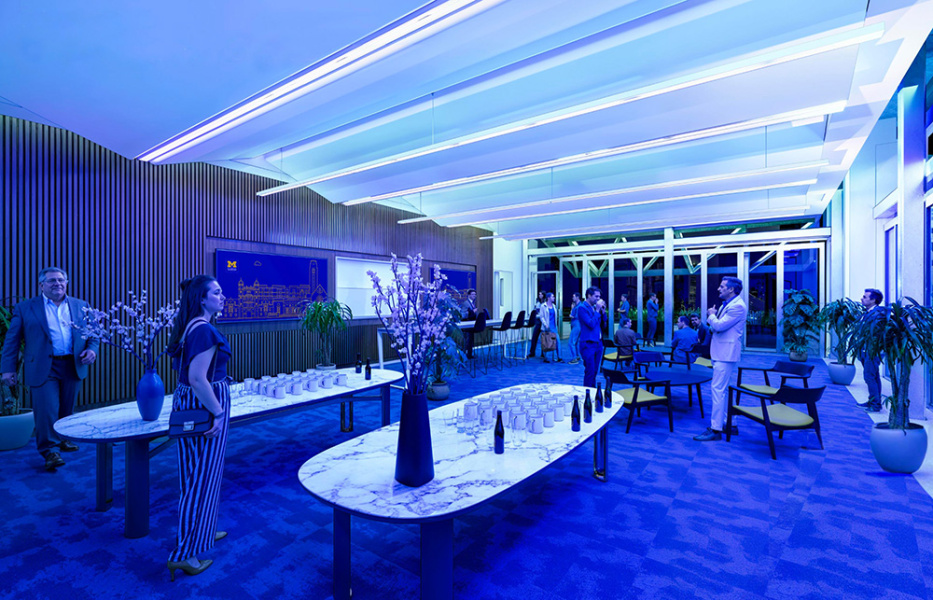 A modern, blue-lit lounge features elegant marble tables set for a reception with bottles and glasses. People mingle, creating a lively yet sophisticated atmosphere.