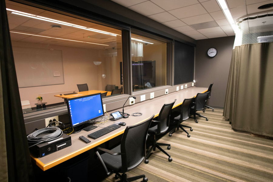 Observation room with multiple chairs, computers, and a large window looking into an adjacent meeting room.