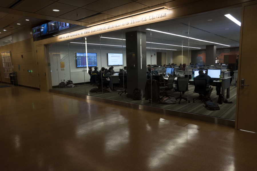 Students work at computer desks in a modern classroom with glass walls and overhead monitors.