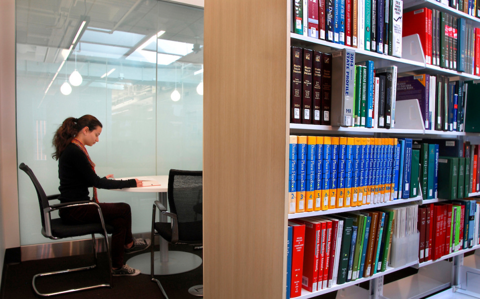 Person studying alone at a desk in a glass-walled library study room beside bookshelves.