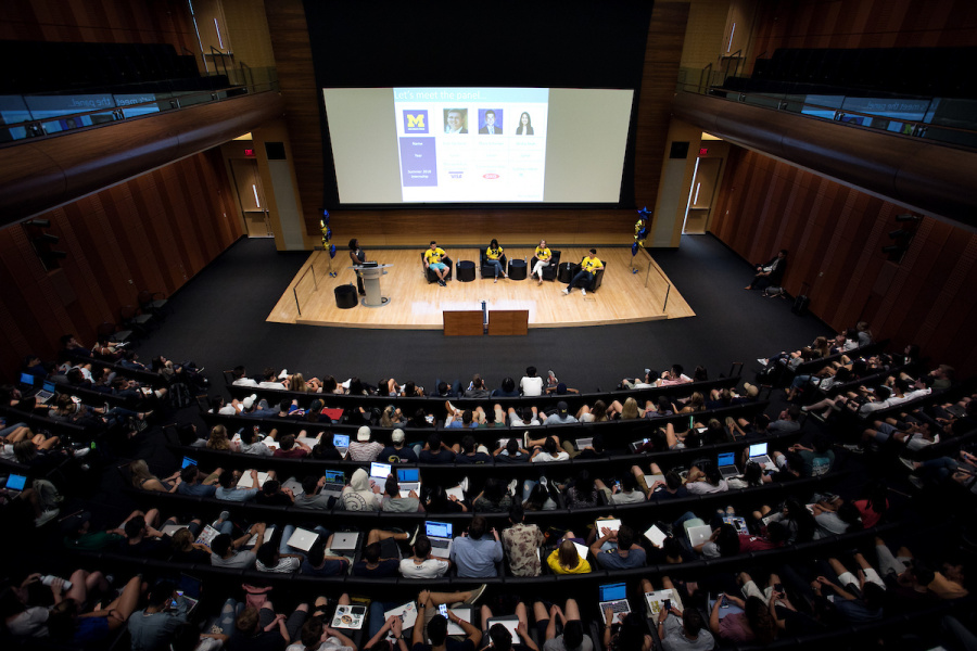 Large audience attends a conference in an auditorium with a presentation projected on a big screen.