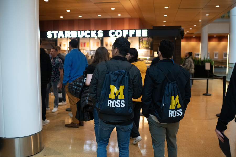 People with "M ROSS" backpacks wait in line at a Starbucks coffee shop.