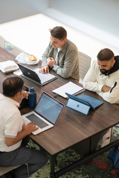 Three people working on laptops around a conference table with notebooks and food containers.