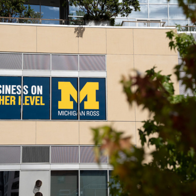 The image shows a beige building facade with a large sign reading "BUSINESS ON ANOTHER LEVEL" and a prominent yellow "M" for Michigan Ross. Trees frame the view.