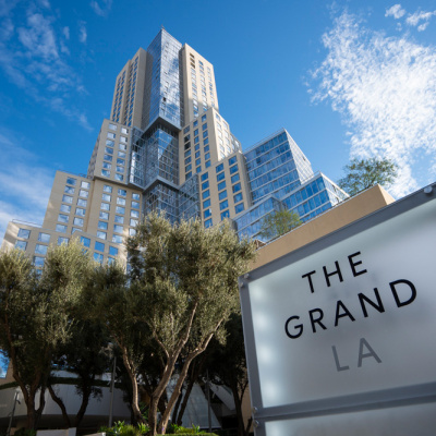 Modern, multi-tiered skyscraper under a blue sky with scattered clouds, framed by trees. "The Grand LA" sign in the foreground conveys elegance.