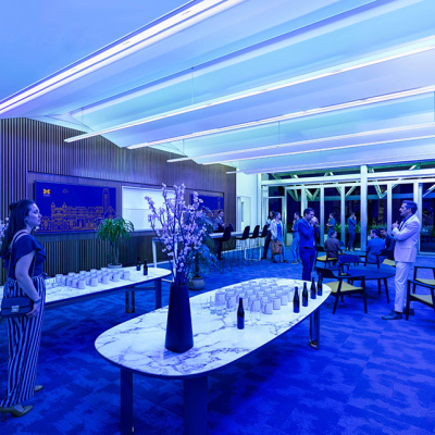 A modern, blue-lit lounge features elegant marble tables set for a reception with bottles and glasses. People mingle, creating a lively yet sophisticated atmosphere.
