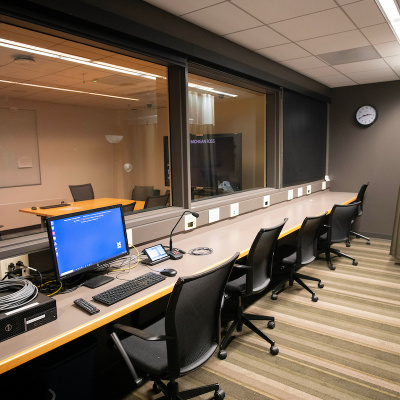Observation room with multiple chairs, computers, and a large window looking into an adjacent meeting room.