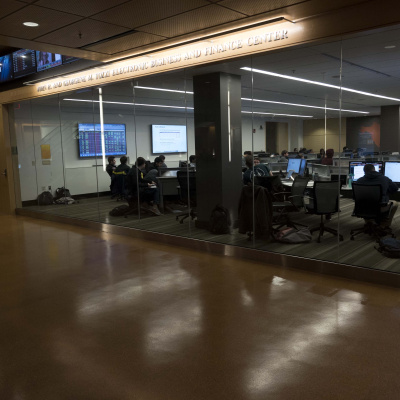 Students work at computer desks in a modern classroom with glass walls and overhead monitors.