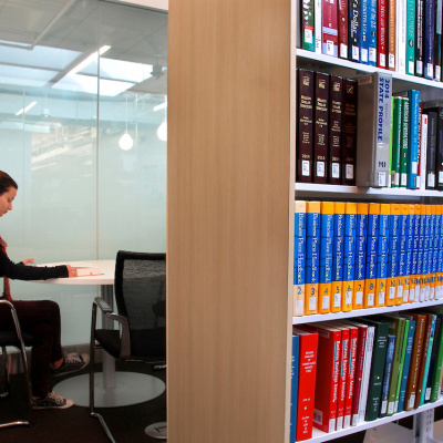 Person studying alone at a desk in a glass-walled library study room beside bookshelves.