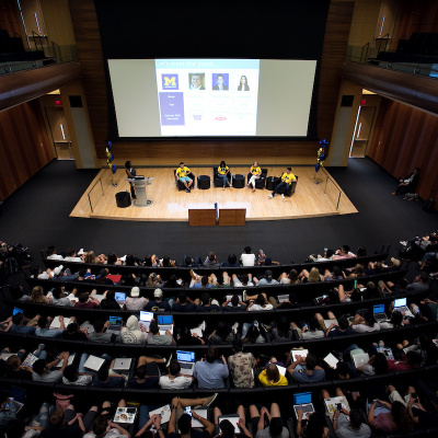Large audience attends a conference in an auditorium with a presentation projected on a big screen.