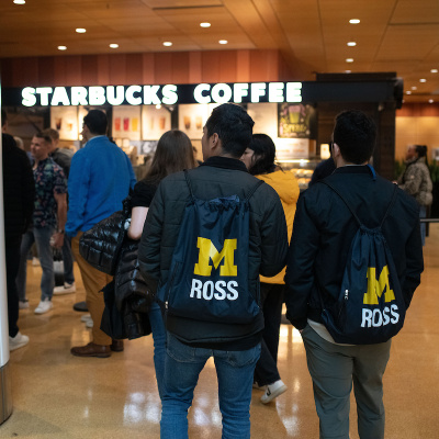People with "M ROSS" backpacks wait in line at a Starbucks coffee shop.