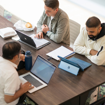 Three people working on laptops around a conference table with notebooks and food containers.