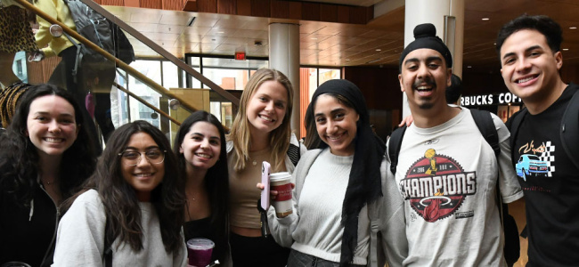 A group of seven friends poses together in a brightly lit room, smiling and holding drinks, with a wooden staircase in the background.