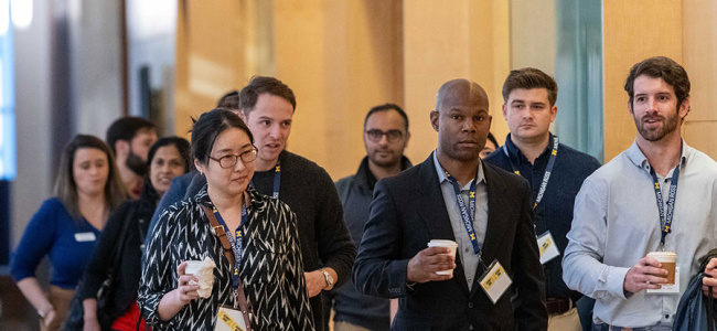 A group of people walking indoors, wearing name badges and holding coffee cups, with a modern backdrop of wood and glass.