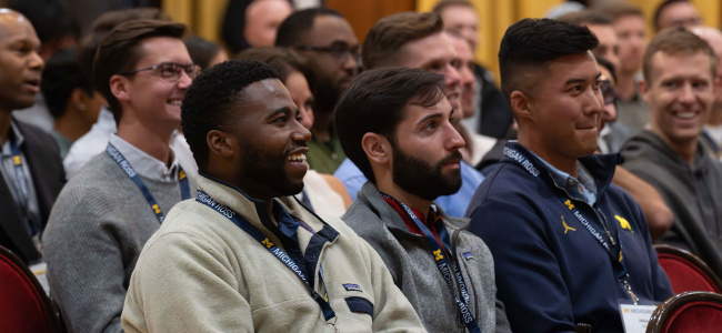 A diverse group of adults sits in a crowded conference room, smiling and attentively listening. They wear casual clothing and name tags, creating a lively, engaged atmosphere.