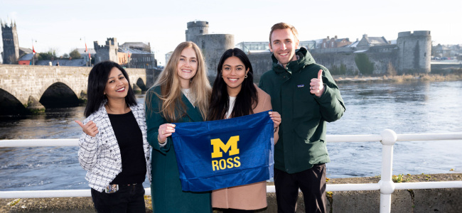 Four people stand by a river, smiling and holding a "Ross" flag with a yellow 'M'. A stone bridge and cityscape are visible in the background.