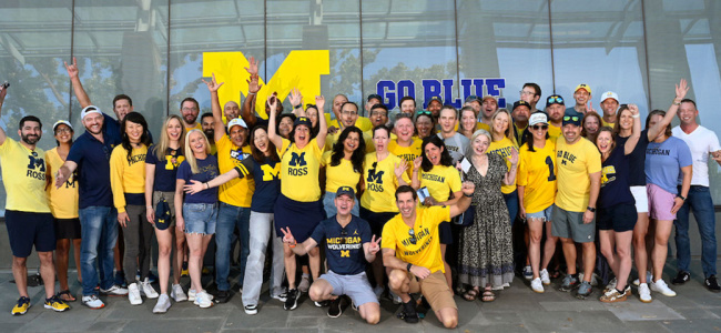 big crowd of people posing in front of window art featuring block M and "Go Blue"