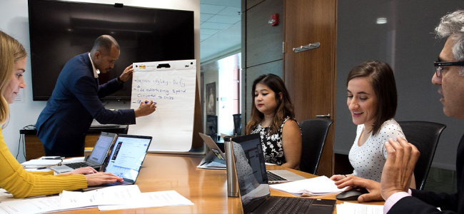 A diverse group of five professionals engaged in a meeting in a modern office. One writes on a flip chart while others work on laptops, appearing focused.