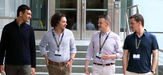 Four men are walking side by side outdoors, wearing ID badges and casual business attire, smiling and conversing, creating a friendly, collaborative atmosphere.